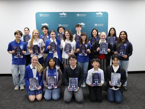 A group of students pose in front of a media wall. They are an ethnically diverse group. They are holding certificates for their participation in the program.