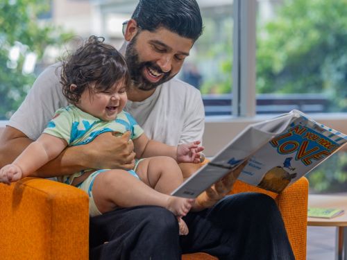 Picture of a man and his baby reading a book in the library.