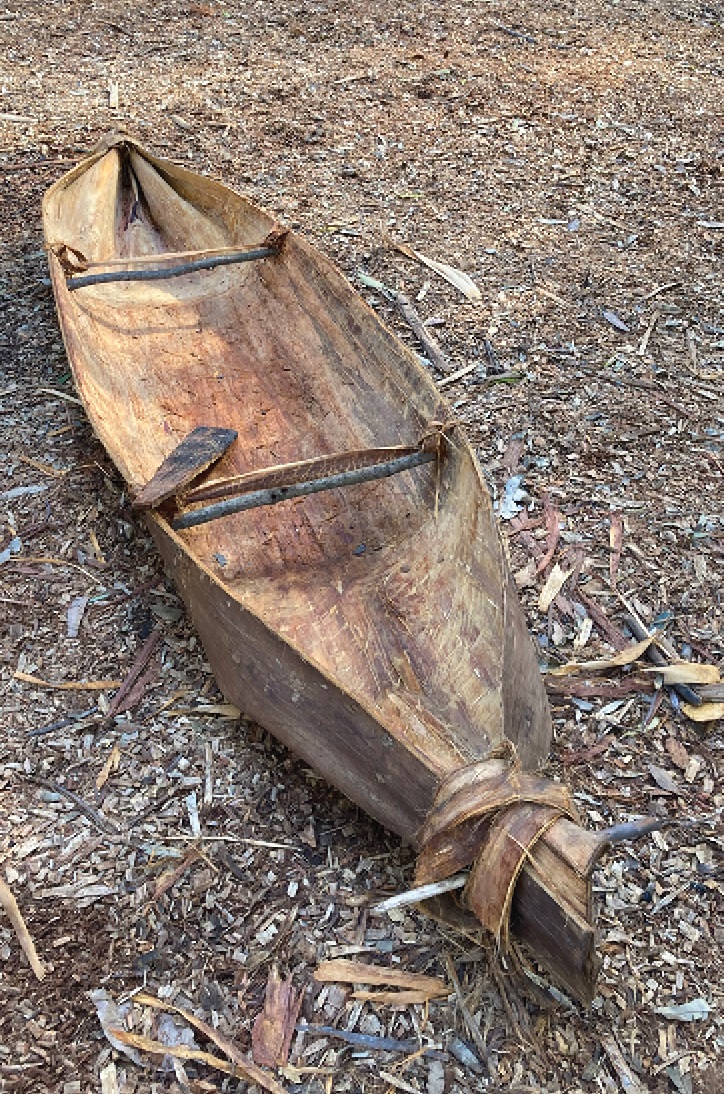 Traditional tied bark canoe with a wooden paddle, placed on the ground.