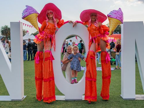 Mother and daughter peering through the giant O in NOM, with two brightly-dressed women on stilts holding giant ice cream cut out stand either side