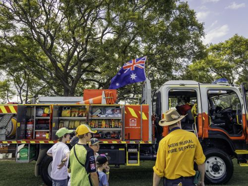 An image of a Rural  Fire Service struck parked in a field. Thee young children are looking at it and standing next to an RFS volunteer